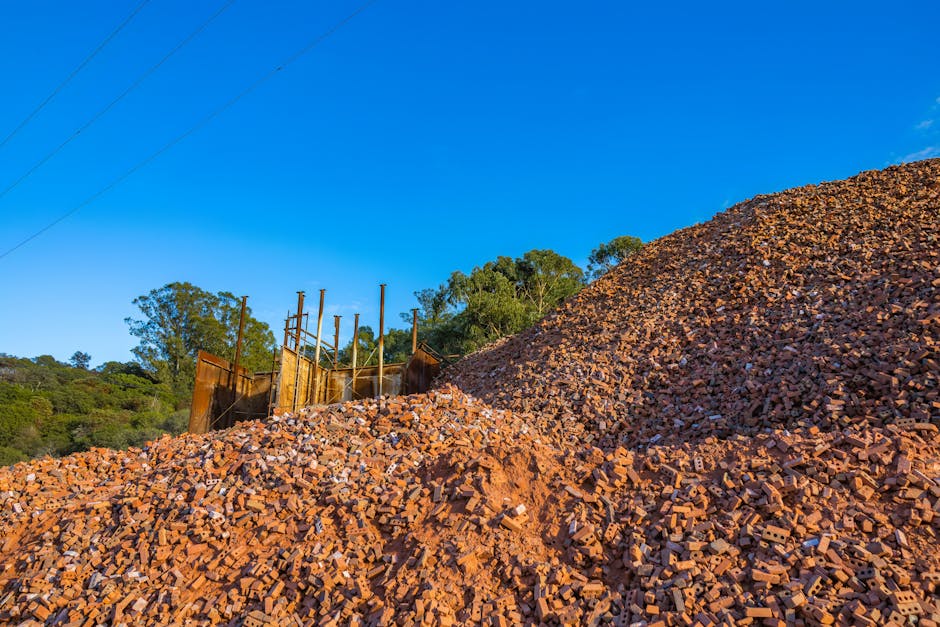 A large pile of red bricks and construction debris, consisting of broken bricks and fragments with rough, uneven textures, dominates the foreground of the image. The debris forms a mound that extends across most of the lower half of the scene, with some scattered smaller pieces visible at its base. Behind the pile, an aged, rusted metal construction fence or barrier, supported by thin metal poles, stands partially obscured by the pile. In the background, there is greenery with tall trees and shrubs, indicating a semi-rural or suburban environment. The sky above is clear and bright blue, with a few faint power lines visible in the upper left corner, adding to the natural yet urban context. The scene appears to be an outdoor site, possibly related to building demolition or renovation waste, which aligns with the scope of independent rubbish clearance services offered by Waste Clearance Mayfair.