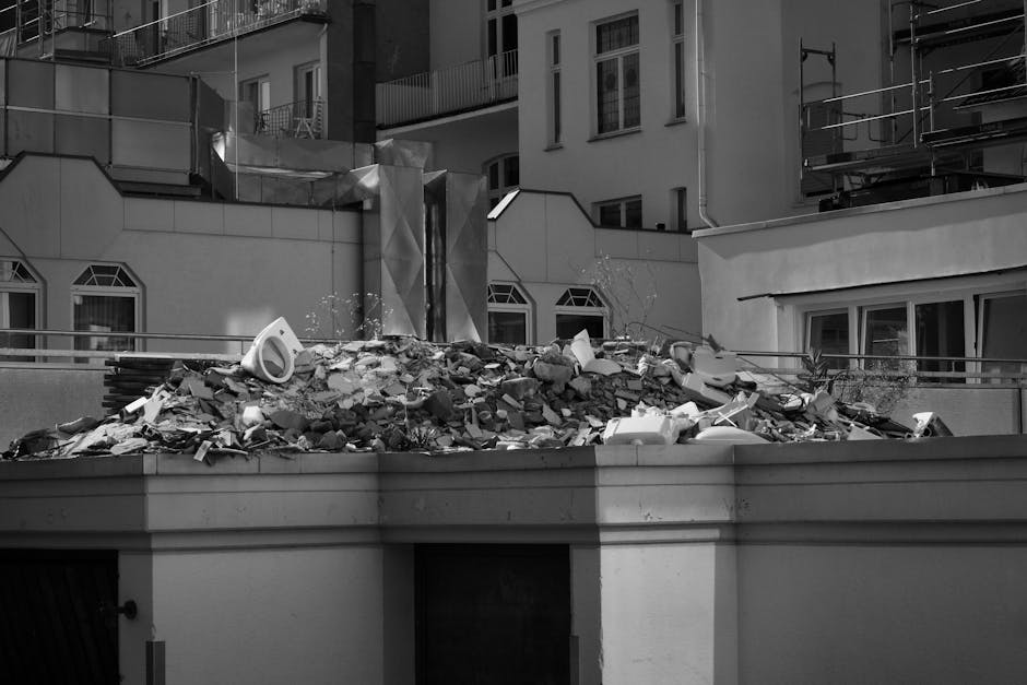 A large pile of construction or demolition debris is accumulated on a flat rooftop or terrace area of a building in an urban setting. The debris includes broken concrete, bricks, pieces of drywall, and discarded plastic items such as pipes and packaging materials, exhibiting a rough, uneven texture with jagged edges and a mix of lighter and darker grey tones. The pile is situated towards the center of the image, with some debris spilling over the edges, and appears to be in the process of being cleared or temporarily stored before removal. Surrounding the debris are metal railings and the facades of nearby buildings with a mix of modern and traditional architectural features, including several windows with geometric framing. The environment suggests an active construction or renovation site in a city area, where private waste management services like those provided by Waste Clearance Mayfair might handle the removal of such bulky waste materials from rooftops or similar hard-to-access locations. The overall atmosphere is well-lit, with natural daylight illuminating the scene and casting soft shadows. The scene underscores the importance of professional rubbish removal services specializing in off-site clearance of construction debris in densely populated urban areas.
