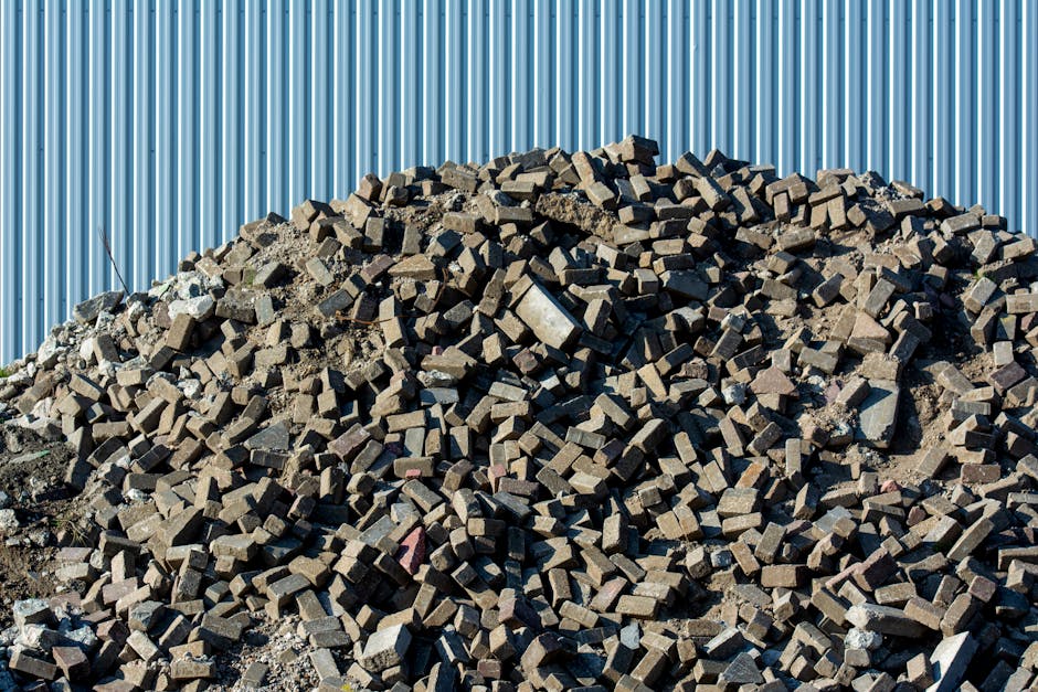 A large pile of red bricks and construction debris, consisting of broken bricks and fragments with rough, uneven textures, dominates the foreground of the image. The debris forms a mound that extends across most of the lower half of the scene, with some scattered smaller pieces visible at its base. Behind the pile, an aged, rusted metal construction fence or barrier, supported by thin metal poles, stands partially obscured by the pile. In the background, there is greenery with tall trees and shrubs, indicating a semi-rural or suburban environment. The sky above is clear and bright blue, with a few faint power lines visible in the upper left corner, adding to the natural yet urban context. The scene appears to be an outdoor site, possibly related to building demolition or renovation waste, which aligns with the scope of independent rubbish clearance services offered by Waste Clearance Mayfair.