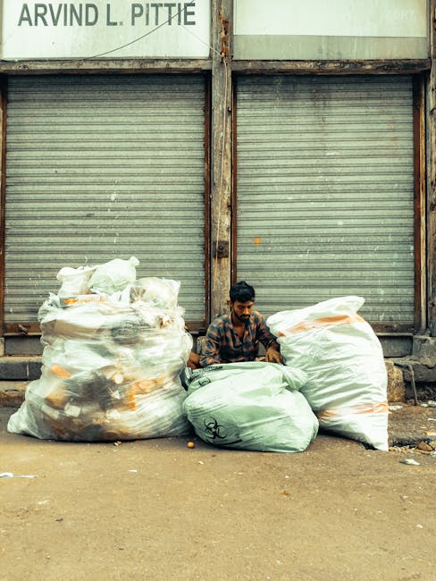 A man with dark hair and a patterned shirt is seated on the ground in front of a closed, weathered metal shutter with horizontal ridges, which appears to be part of an industrial or commercial building. He is surrounded by multiple large rubbish bags, some white and others green, filled with discarded items. The bags are stacked and leaning against each other, occupying most of the foreground. The surface underneath is concrete, showing some dirt and debris. Behind the man, the metal shutter extends vertically, with rust and discoloration visible along the edges. Above the shutter, part of a sign with partially visible text can be seen, suggesting an urban or commercial environment typical of an area where private rubbish collection or waste management services, such as those provided by Waste Clearance Mayfair, operate. The overall scene conveys an off-street waste collection scenario aligned with independent or alternative rubbish removal methods commonly used in urban settings.