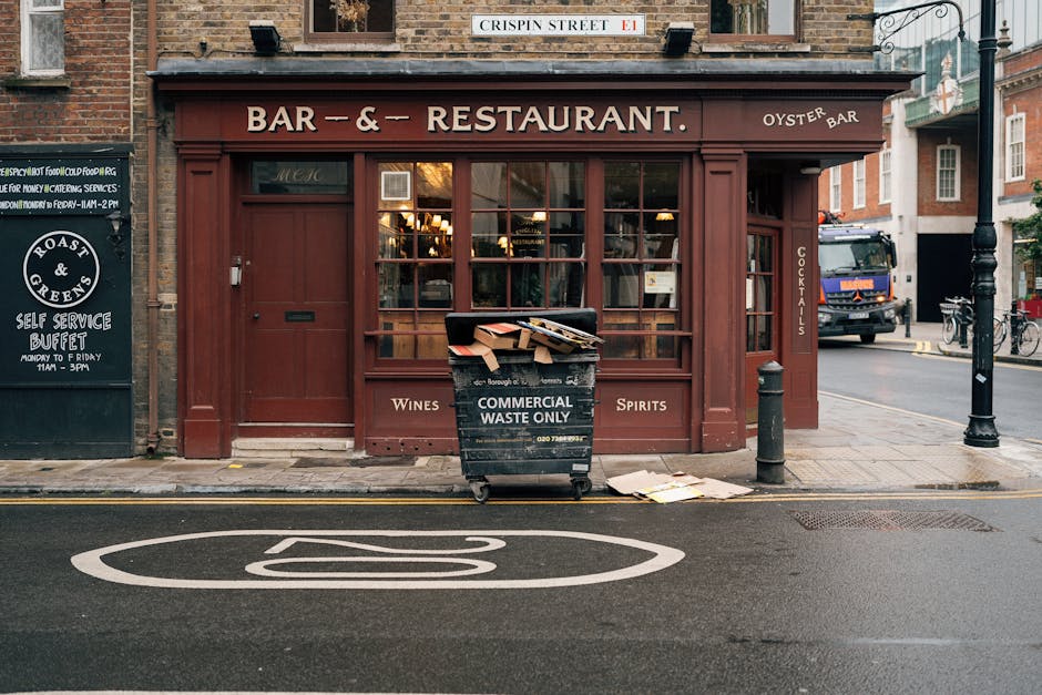 A black wheeled waste collection bin labeled 'Commercial Waste Only' positioned on the paved pavement outside a restaurant, containing assorted flattened cardboard boxes and paper waste with some pieces spilling onto the sidewalk. The restaurant has wooden-framed large windows and a maroon exterior with signage indicating it serves wines and spirits, as well as offering oyster dishes. The scene includes a nearby black bollard and a black pole with decorative lamp post fixtures on a typical city street, with a parked truck visible in the background. The environment appears to be an urban setting with brick buildings and street markings, suggesting an area where independent waste removal or on-site clearance services may be employed by local businesses, exemplified by Waste Clearance Mayfair’s operational context.