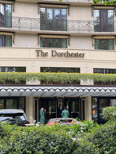 The exterior of The Dorchester hotel in Mayfair, featuring its cream-colored stone facade with elegant decorative trim. The hotel's name is prominently displayed in large, brown lettering above the entrance. A neatly maintained hedge runs along the front of the building, partially obscuring the ground floor windows. Two men in formal business attire are standing near the black entrance doors, engaging in conversation, while another person in a green suit is nearby. A black vehicle is parked close to the entrance on the left side, and another darker vehicle is visible on the right. The upper floors showcase tall windows framed by black railing balconies, with some glass doors open to small outdoor terraces. The scene is set during daylight with natural lighting, giving a clear view of the hotel’s architectural details and surrounding greenery, in a context where private property and high-end hospitality intersect with the local environment, relevant to independent waste management services in the area.