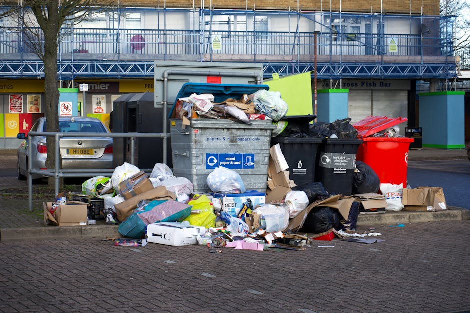 A black wheeled waste collection bin labeled 'Commercial Waste Only' positioned on the paved pavement outside a restaurant, containing assorted flattened cardboard boxes and paper waste with some pieces spilling onto the sidewalk. The restaurant has wooden-framed large windows and a maroon exterior with signage indicating it serves wines and spirits, as well as offering oyster dishes. The scene includes a nearby black bollard and a black pole with decorative lamp post fixtures on a typical city street, with a parked truck visible in the background. The environment appears to be an urban setting with brick buildings and street markings, suggesting an area where independent waste removal or on-site clearance services may be employed by local businesses, exemplified by Waste Clearance Mayfair’s operational context.
