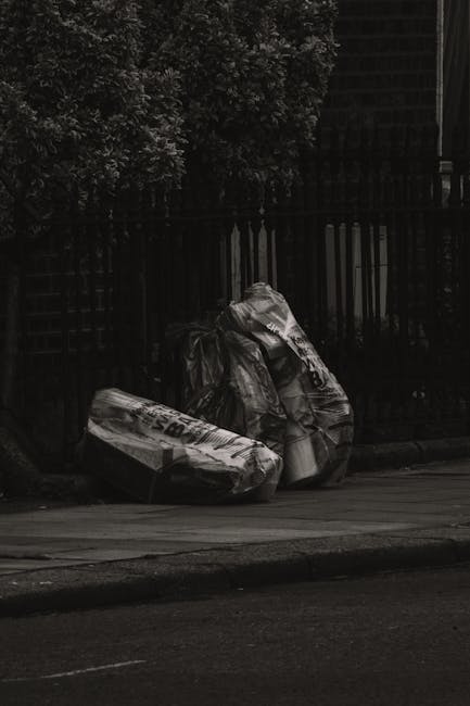 Two large black plastic rubbish bags, slightly crumpled, are placed on a sidewalk near a curb. The bags appear to be filled with waste material and are positioned close together, leaning slightly against a dark metal fence with vertical bars. Behind the fence, dense foliage of trees or shrubs is visible, indicating an outdoor environment in an urban or suburban setting. The scene is captured in low natural light, creating a shadowed and subdued atmosphere. The image suggests an instance of external waste disposal, which could be associated with independent rubbish collection or private waste removal services, such as those offered by Waste Clearance Mayfair, especially as part of an emergency rubbish pickup or same-day disposal arrangement.