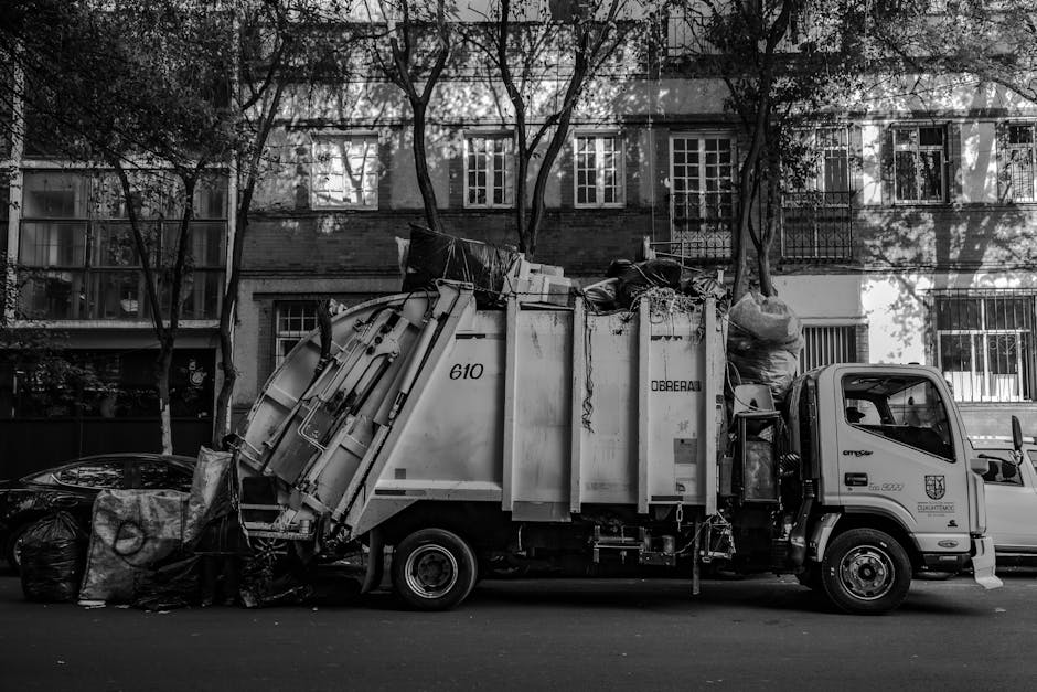 A side view of a commercial waste collection truck parked on a city street in front of residential apartment buildings with multiple floors. The truck is primarily made of metal with a smooth, reflective surface, painted in light shades, and features identifiable equipment for rubbish collection, including a large rear-loading container labeled with the number '610' and some text. The container is filled with mixed waste items, including black garbage bags, cardboard, and other refuse, which appear uncompressed and partially overflowing. The truck's cab is positioned towards the right, with a small window and side mirror visible, and the surrounding environment includes trees with visible branches casting shadows on the buildings and the vehicle. The scene is captured in natural daylight, contributing to clear visibility of the textures and materials, such as the smooth metal surfaces, the crinkled plastic bags, and the brickwork of the residential structure behind. This image illustrates an example of private rubbish collection services, like those offered by Waste Clearance Mayfair, as an alternative method for waste disposal in urban areas.