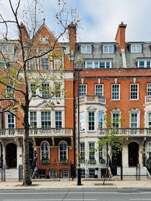 A row of historic red brick and white stucco residential buildings with ornate architectural details, such as decorative balustrades, cornices, and sash windows, situated along a city street. The buildings feature multiple stories, with some having small balconies and bay windows, and are framed by leafless trees that stretch upward, partially obscuring the upper floors. In the foreground, a paved sidewalk runs parallel to the street, with a black-and-yellow traffic bollard and a streetlamp standing between the buildings. The sky above is partly cloudy, with patches of blue visible. This environment highlights the urban context where private waste collection services, like those provided by Waste Clearance Mayfair, may be employed for on-site clearance or rubbish removal from such properties, supporting alternative waste management solutions in the area.