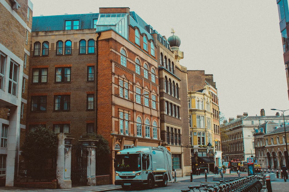 A street scene featuring a range of historic and modern buildings with varying architectural styles, including brick facades, decorative window frames, and a dome-topped turret. In the foreground, a waste collection vehicle with a grey and blue body is parked on the street, near a bicycle rack and pedestrian pavement. The vehicle is positioned adjacent to a gated entrance with stone pillars, possibly leading to an enclosed area or building entrance. Surrounding the scene are other commercial and residential structures, some with ornate details and large windows. The atmosphere is overcast with diffused natural lighting, and the street is lined with bollards and street furniture, indicating an urban environment that may accommodate private waste collection services by companies such as Waste Clearance Mayfair. The scene emphasizes an area where private rubbish collection and on-site clearance might take place within a city context, supported by the visible waste management vehicle and urban setting.