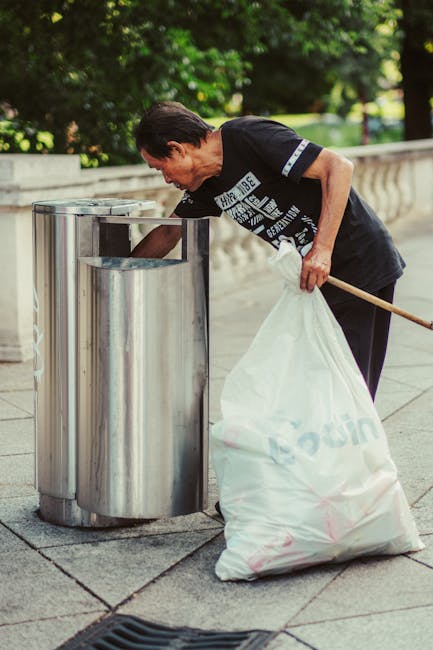 A middle-aged man with dark hair, wearing a black T-shirt with white and blue text, is bending over to dispose of rubbish into a circular stainless steel public trash bin on a paved sidewalk. He is holding a large white plastic bag filled with waste, which appears to contain various items, and using a wooden stick or brush to assist in depositing the rubbish. The background features green leafy trees and a low stone wall, suggesting a park or street setting. The scene is illuminated by natural daylight, highlighting the reflective surface of the metal bin and the texture of the pavement. This image depicts an individual undertaking independent waste disposal, aligning with private rubbish collection or on-site rubbish removal services, as provided by waste management specialists such as Waste Clearance Mayfair.