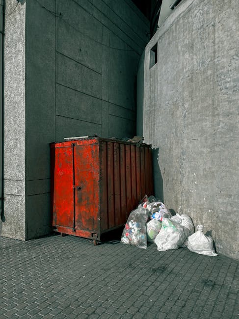 A weathered red metal dumpster with a slightly rusted surface is positioned against a dark green wall and a light grey concrete wall, located on a paved urban sidewalk. The dumpster's surface shows signs of chipping paint and corrosion, indicating exposure to the elements. Next to it, several white plastic trash bags filled with assorted waste are piled nearby, some of which are piled directly on the ground and appear loosely tied with visible contents inside. The environment appears to be an alleyway or service area adjacent to a modern building, with textured tiles on the pavement and a small rectangular opening higher up on the grey wall, possibly for ventilation or access. The lighting is natural, casting soft shadows in the scene, consistent with daylight conditions. This setup suggests an informal waste disposal spot for commercial or residential waste collection, typical of independent rubbish handling or on-site clearance services often undertaken by waste management specialists such as Waste Clearance Mayfair.