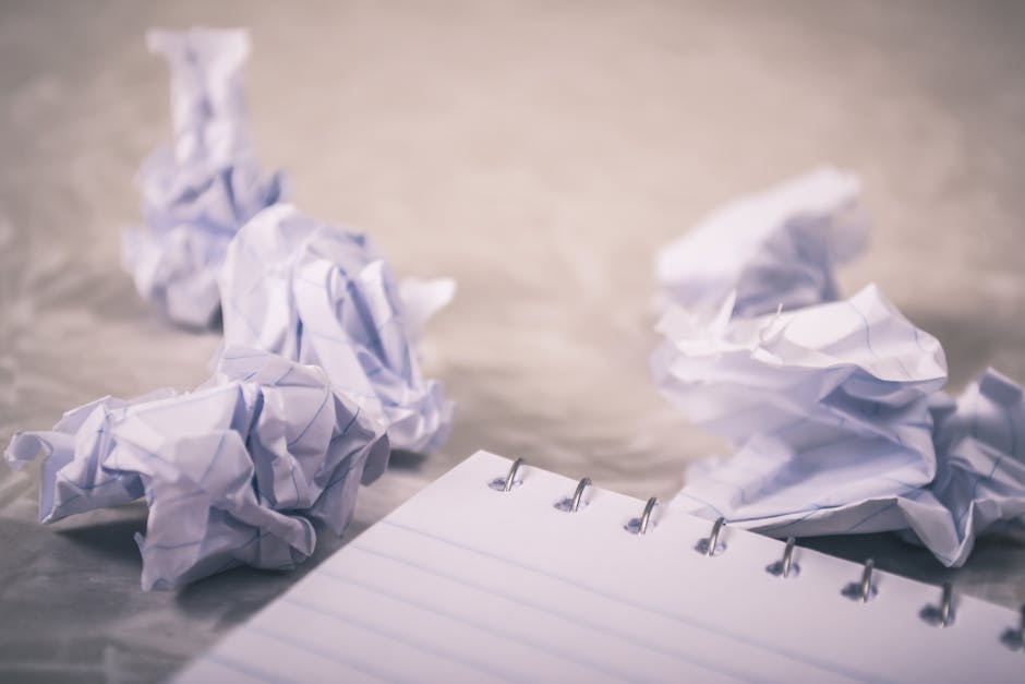 A close-up view of several crumpled sheets of white paper scattered on a smooth, reflective surface, with some pieces overlapping and others lying flat. The paper appears to be standard A4 weight with visible creases and torn edges, suggesting they have been discarded after use. In the foreground, a small open notebook with white lined pages is partially visible, with metal rings binding the pages together. The background is blurred, emphasizing the crumpled paper and the notebook. The lighting is soft and neutral, highlighting the textures of the wrinkled paper and creating subtle shadows that suggest a clean, indoor environment suitable for an interior shot related to clutter or waste removal activities, such as those handled by waste clearance services. This scene visually represents disposed office or paper waste, similar to what might be collected during private rubbish collection or on-site clearance by waste management professionals.