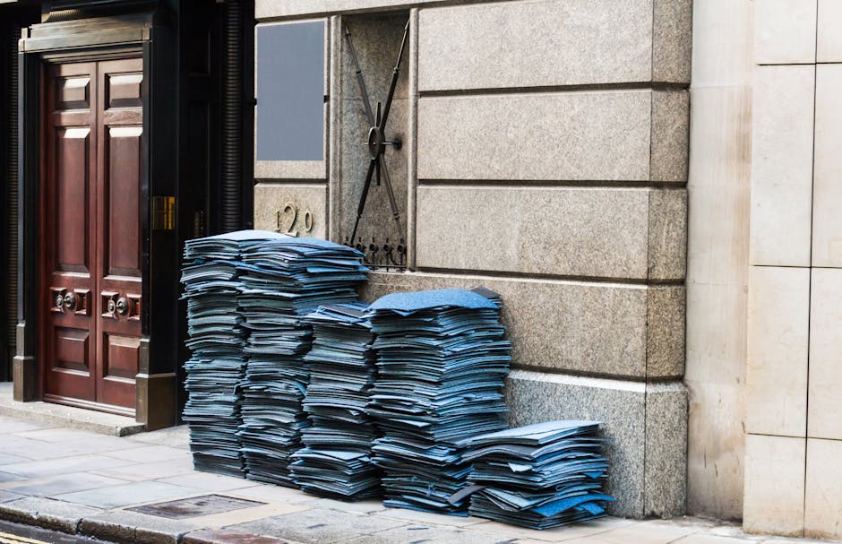 A large stack of blue a4-sized files and documents, piled against a grey stone building exterior on a paved sidewalk. The papers appear disorganized, with some slightly crumpled or unevenly stacked. To the left, there is a dark wooden door with metallic handles, framed by black trim, and adjacent to a metal security shutter that is partially open. On the wall behind the stacks, there is a metal mailbox or utility box, with a lock and a small handle, attached to the building. The environment looks like an urban setting, potentially indicating a private or commercial property, where such a collection of office or legal documents might be awaiting collection or disposal. The scene may relate to independent rubbish removal or on-site clearance involving document waste, with Waste Clearance Mayfair capable of handling such rubbish efficiently and professionally, supporting alternative waste disposal needs outside local authority services.