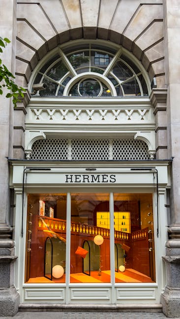 A storefront with a classic architectural facade featuring a large semi-circular arched window with multiple panes set in a stone frame above a white-painted wooden shopfront displaying the name 'HERMES' on a signboard. Inside the store, visible through large glass windows, are warm lighting fixtures, a staircase with a decorative wooden balustrade, and various display objects, including illuminated boxes and circular decorative elements. The building's exterior is constructed from light-colored stone, with detailed molding and a surrounding urban environment. The scene captures the elegant storefront design typical of high-end retail locations, with a focus on clarity and detail relevant to private retail waste management and on-site clearance considerations, corresponding with waste clearance services in Mayfair.