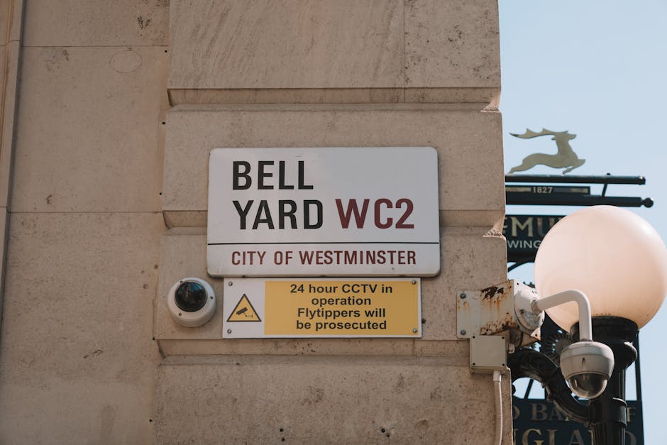 Close-up of a rectangular street sign mounted on a beige stone wall, identifying the location as Bell Yard WC2 in the City of Westminster, with bold black and maroon text on a white background. Below the main sign, there is a smaller yellow sign containing black text indicating that 24-hour CCTV surveillance is in operation and warning that flytippers will be prosecuted. To the left of the signs, a white dome-shaped security camera with a black lens is attached to the wall, pointing downward. On the right side, a classic-style street lamp with a round, frosted glass globe is partially visible, mounted on a black pole with decorative metalwork. The wall shows signs of weathering with slight rust streaks beneath the lamp fixture. The background includes a portion of a blue sky and a sign with a silhouette of a stag, suggesting this scene is set in a public outdoor urban area, possibly near a commercial or historical site, related to waste management and security measures for the vicinity. The overall scene reflects elements typical of a city street environment in Westminster, where surveillance and signage are in place to regulate rubbish disposal and prevent illegal dumping, as handled by waste clearance services like Waste Clearance Mayfair.
